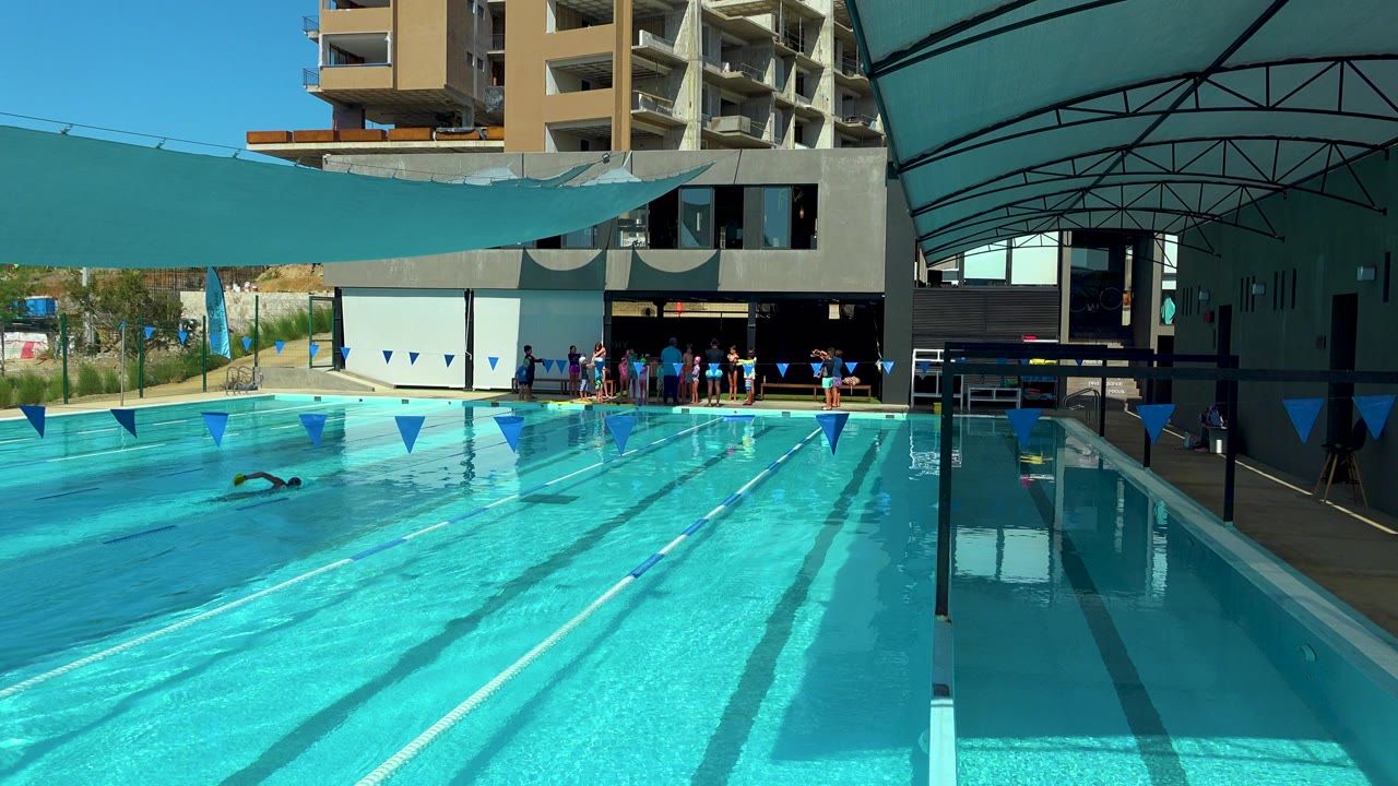 Cabo San Lucas hotel lap pool with bright blue water, a swimmer at left, blue lane flags across the lanes, and people gathered near the shaded deck.