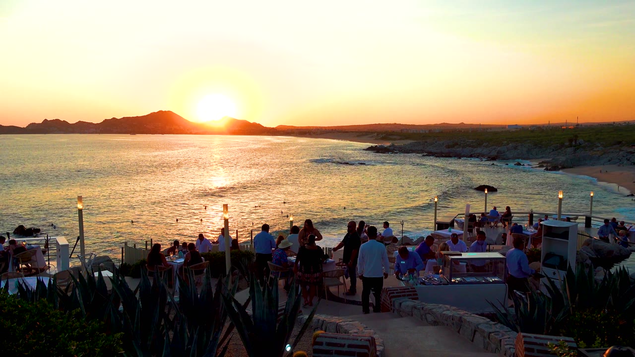 Cabo San Lucas terrace scene with people seated near the railing, a bright sun near the hills, small waves below, and dark plants in the foreground.