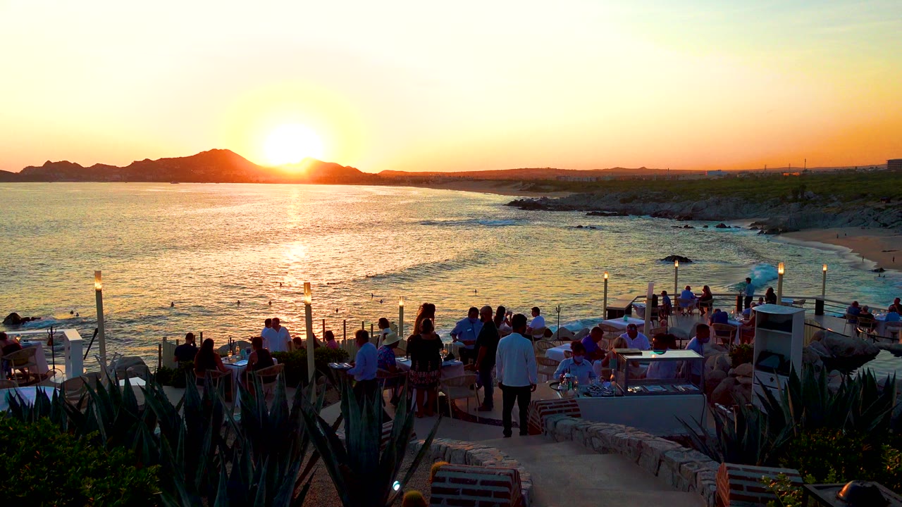 Wide view in Cabo San Lucas, Mexico, with people on an oceanfront terrace as the sun rises near dark mountains and light reflects on the water.