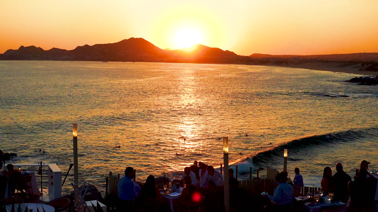 In Cabo San Lucas, Mexico, the sun rises above a dark mountain ridge while people sit and stand in silhouette on an oceanfront terrace below.