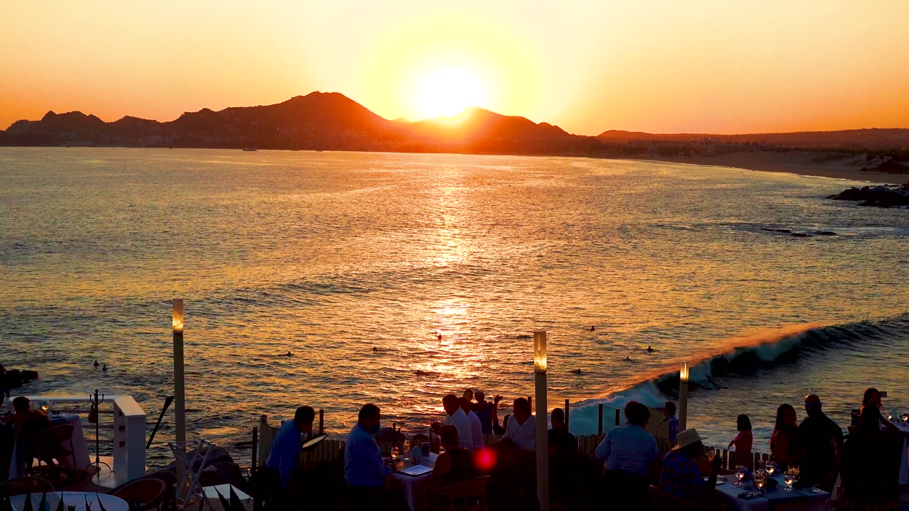 A curling wave forms below the terrace as the sun hangs above the mountain line and silhouetted guests face the ocean in Cabo San Lucas.