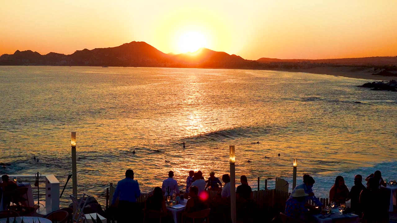Cabo San Lucas sunset over the ocean, with bright orange reflection on the water and silhouetted people seated at terrace tables in the foreground.