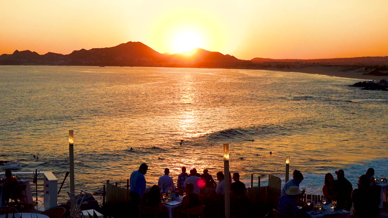 Cabo San Lucas sunset with golden water, dark mountain shapes on the horizon, and foreground diners silhouetted beside lit terrace posts.