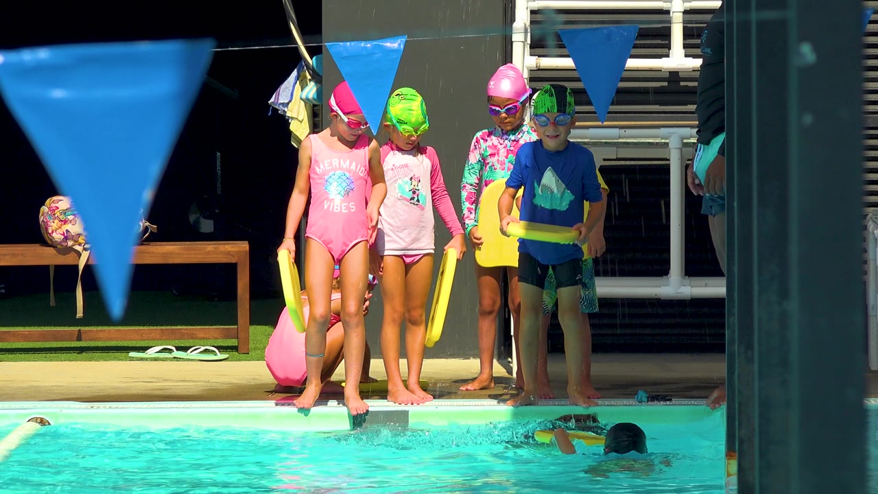 In Cabo San Lucas, a larger group of children with swim caps lines the pool edge as two swimmers appear low in the turquoise water and yellow kickboards are held at the side.