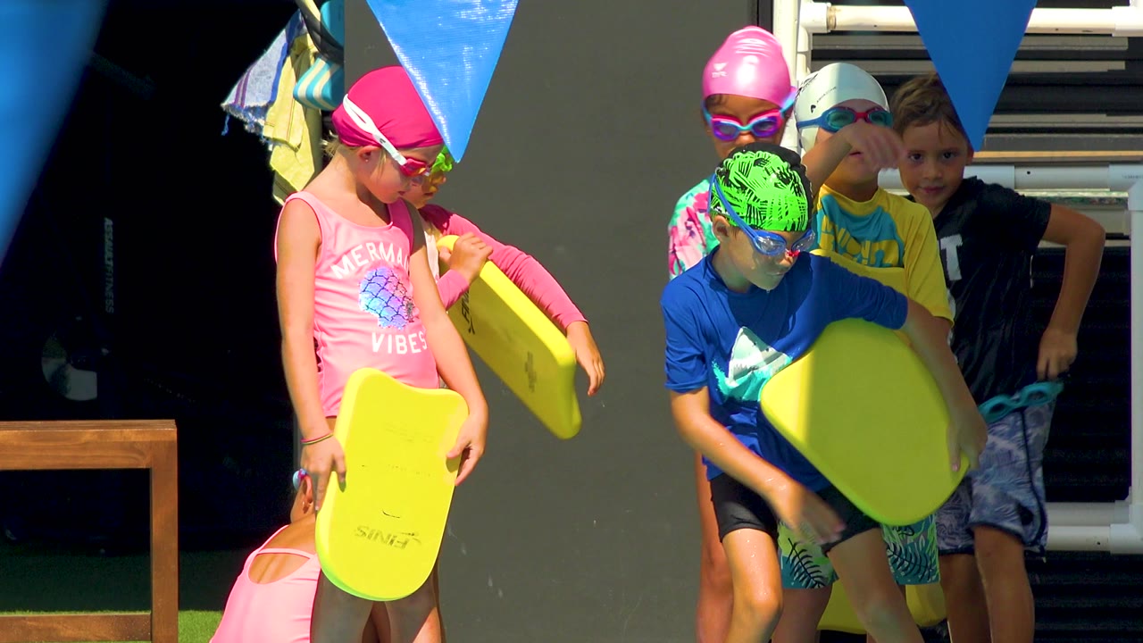 Six children in swim gear gather with yellow kickboards at a sunlit pool edge in Cabo San Lucas, Mexico, with blue lane flags overhead.