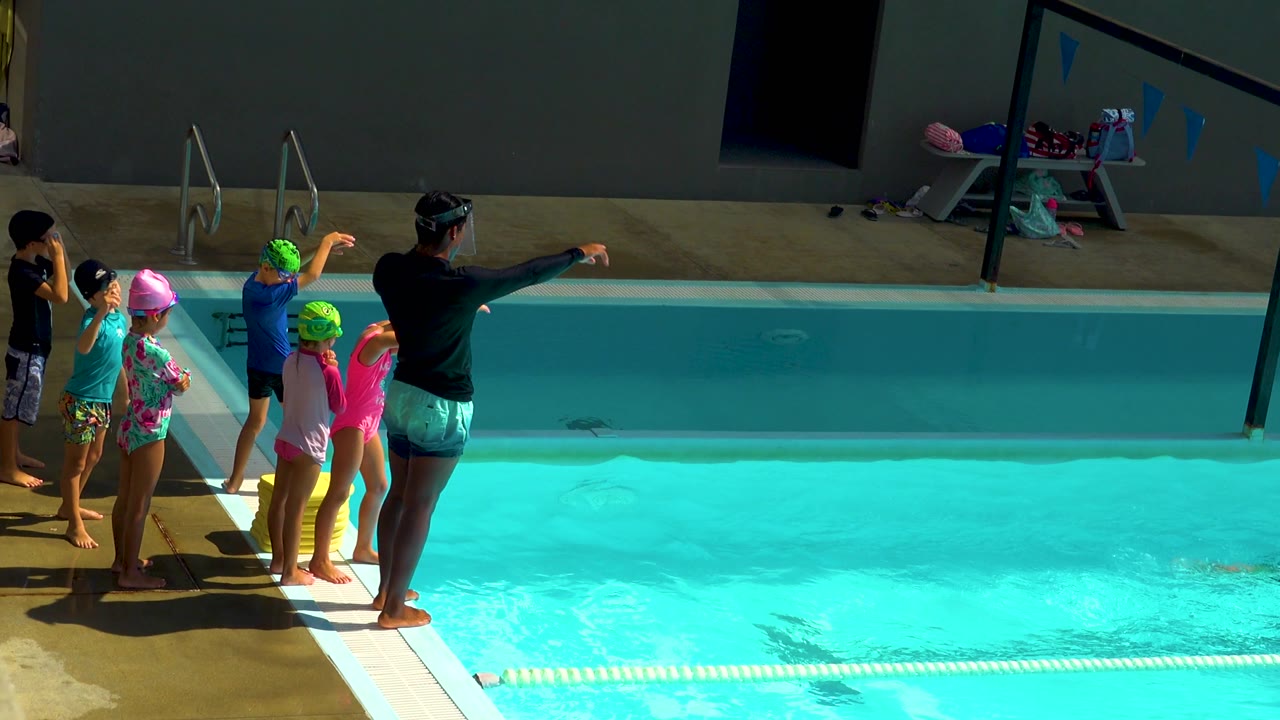 Children in swim caps face the pool while a coach points across the water.