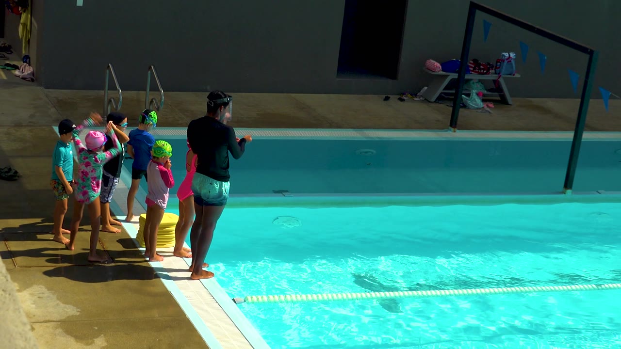 At 0:02, the coach stands closer to the pool edge while children in colorful swim gear line up beside the bright turquoise water.
