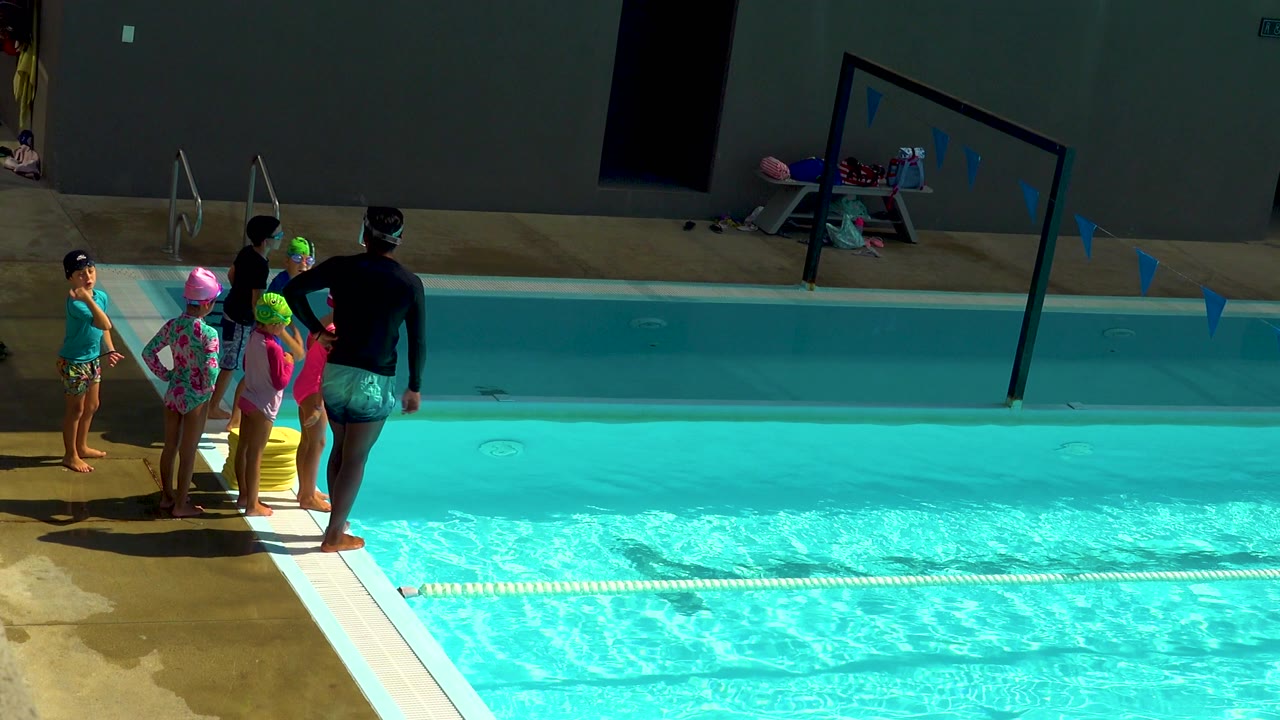 In Cabo San Lucas, Mexico, a swim instructor stands beside several children in swim caps at the pool edge as bright blue water fills the right side of the frame.