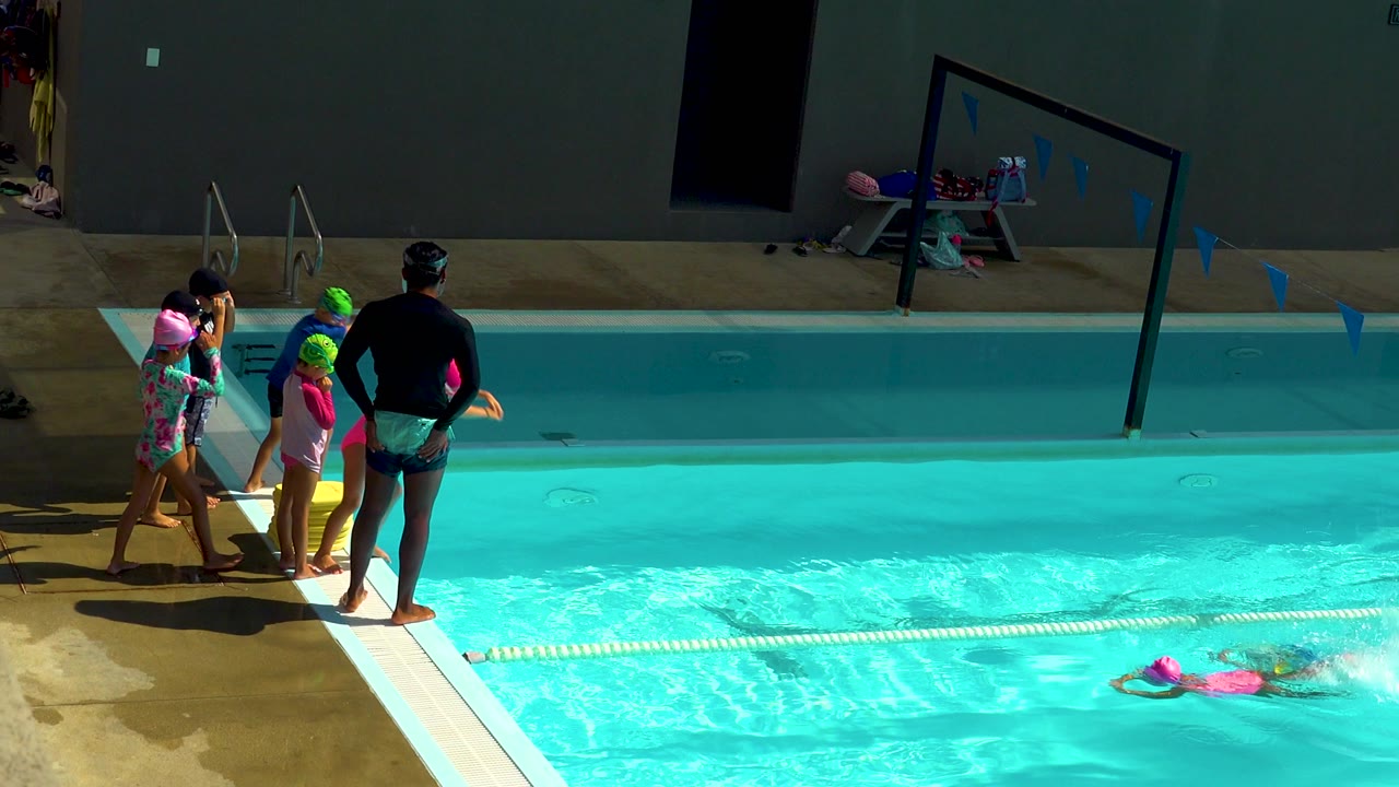 At a Cabo San Lucas pool, children in bright swim caps wait near an adult instructor while two swimmers move through turquoise water beside a lane rope.