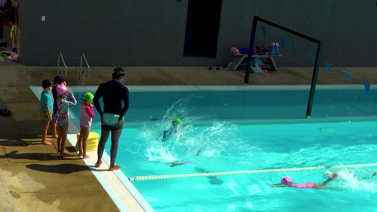 A child in a green cap has just entered the Cabo San Lucas pool, sending up a large splash while other children and an adult stand close to the edge.