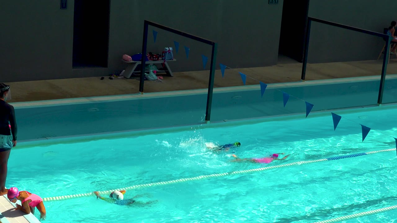 At 0:00 in Cabo San Lucas, Mexico, children swim through turquoise pool lanes while blue flags hang overhead and adults are visible near the pool deck.