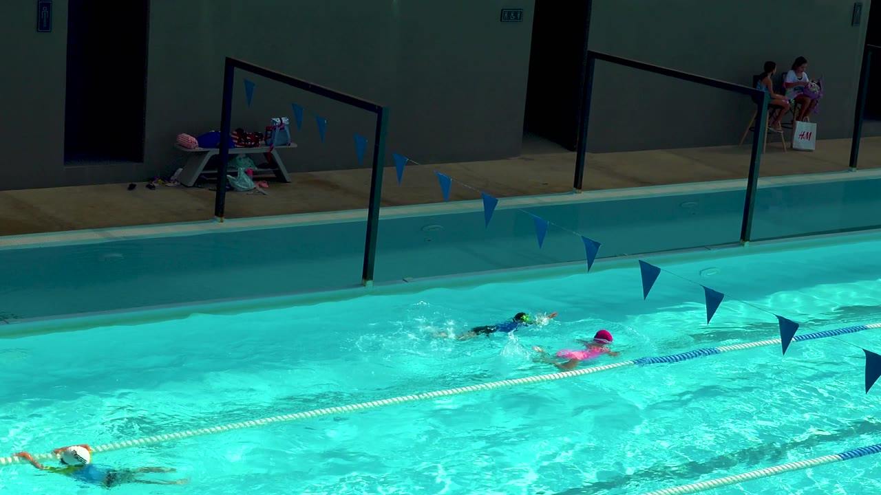 Children swim in bright pool lanes with seated people in the background.