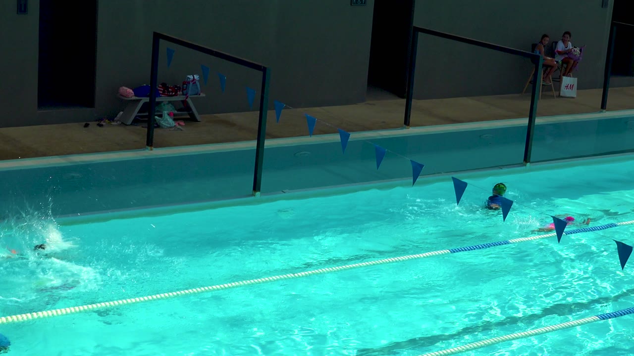 Children swim across a bright pool in Cabo San Lucas, Mexico, with lane ropes, blue pennants, a pool deck, towels, toys, and seated people in the background.