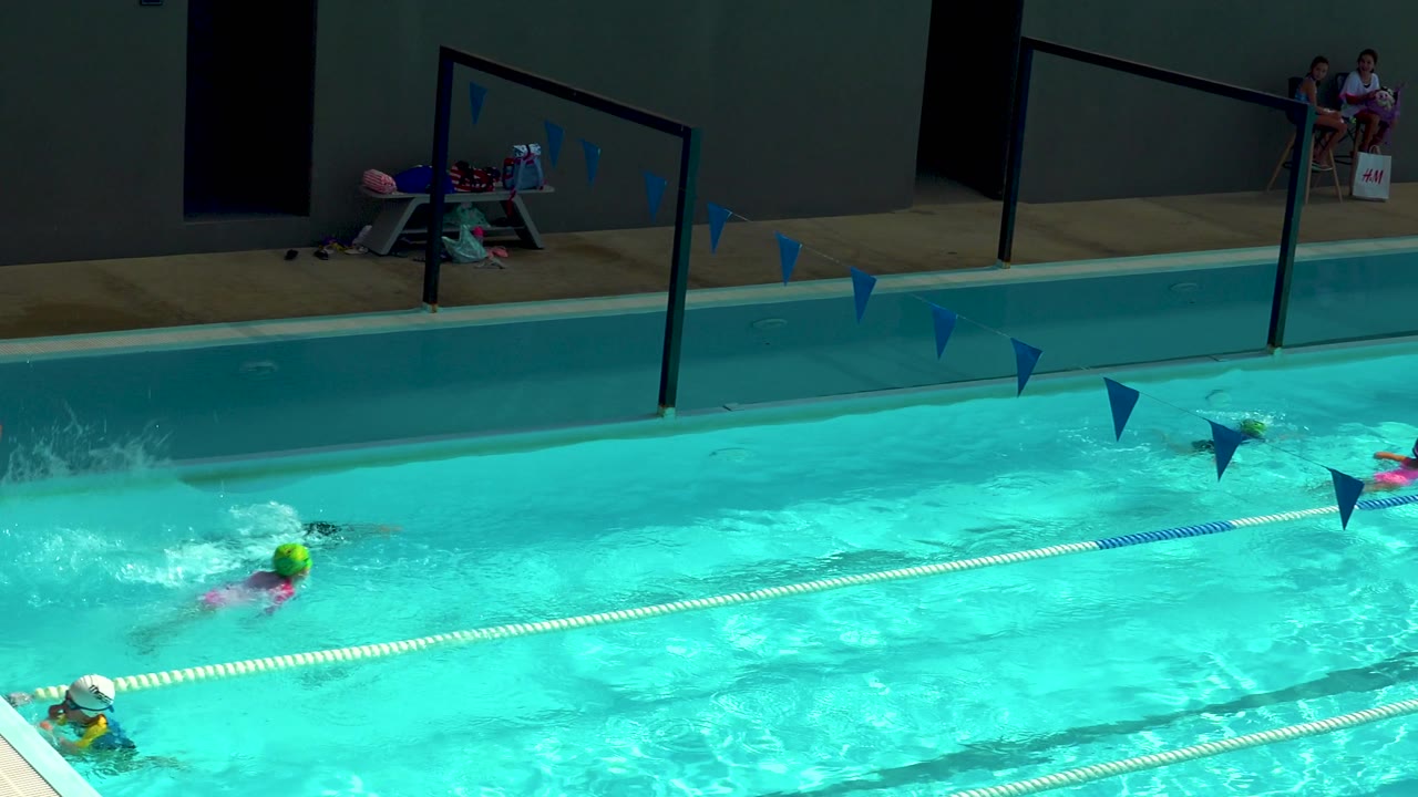 Children continue swimming under blue pennants in the Cabo San Lucas pool, with bright turquoise water, lane ropes, deck railings, and pool gear visible behind them.