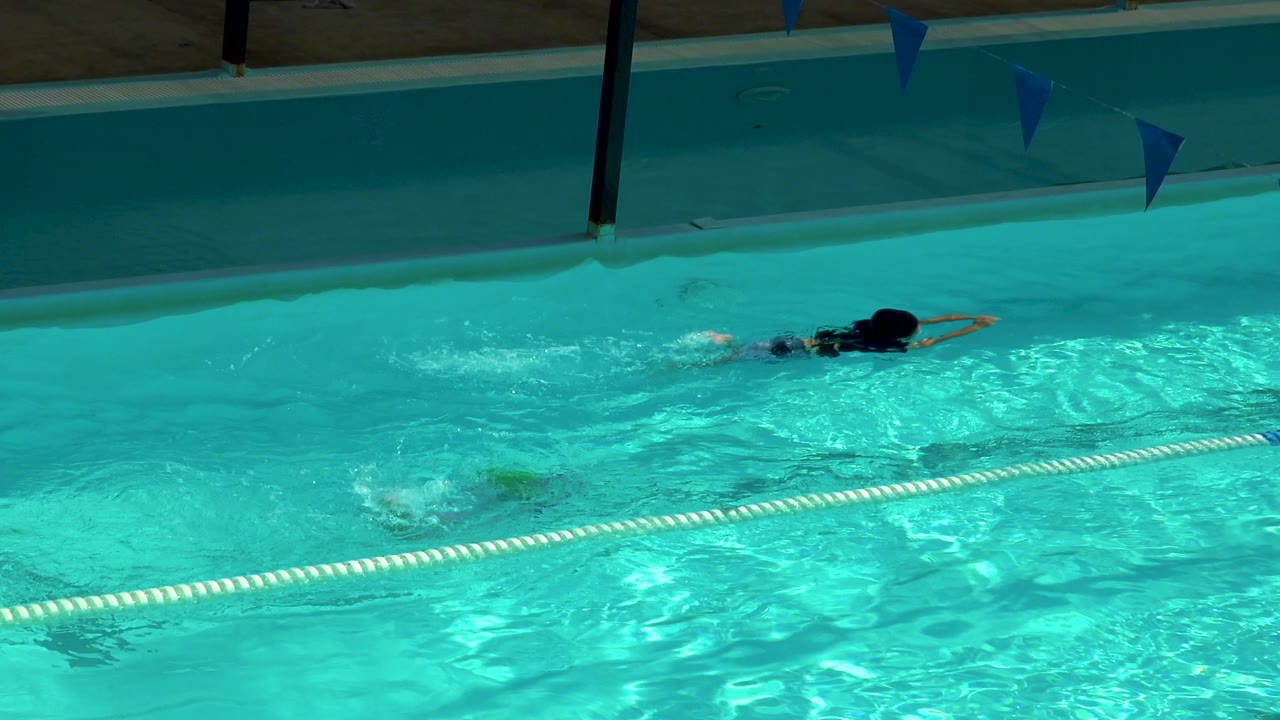 A child swims underwater in a turquoise pool lane in Cabo San Lucas, Mexico, with a second swimmer farther back and blue pennants above the water.