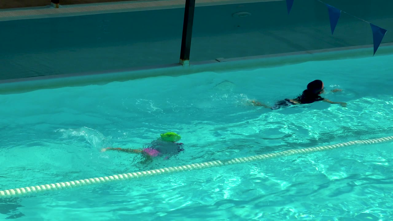 A child in a bright green swim cap and pink top swims beside the lane rope as another swimmer moves ahead in the adjacent lane at the Cabo San Lucas pool.