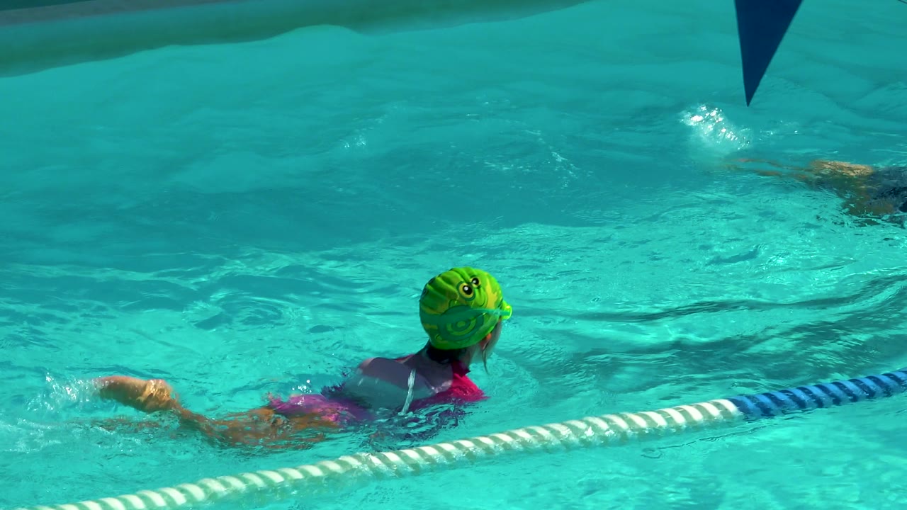 A child in a green swim cap swims through bright turquoise pool water in Cabo San Lucas, Mexico, with a blue-and-white lane rope crossing the foreground.