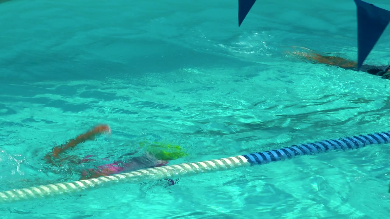 Swimmers are partly submerged in the Cabo San Lucas pool, with blue triangular pennant flags overhead and the lane rope running across the lower frame.