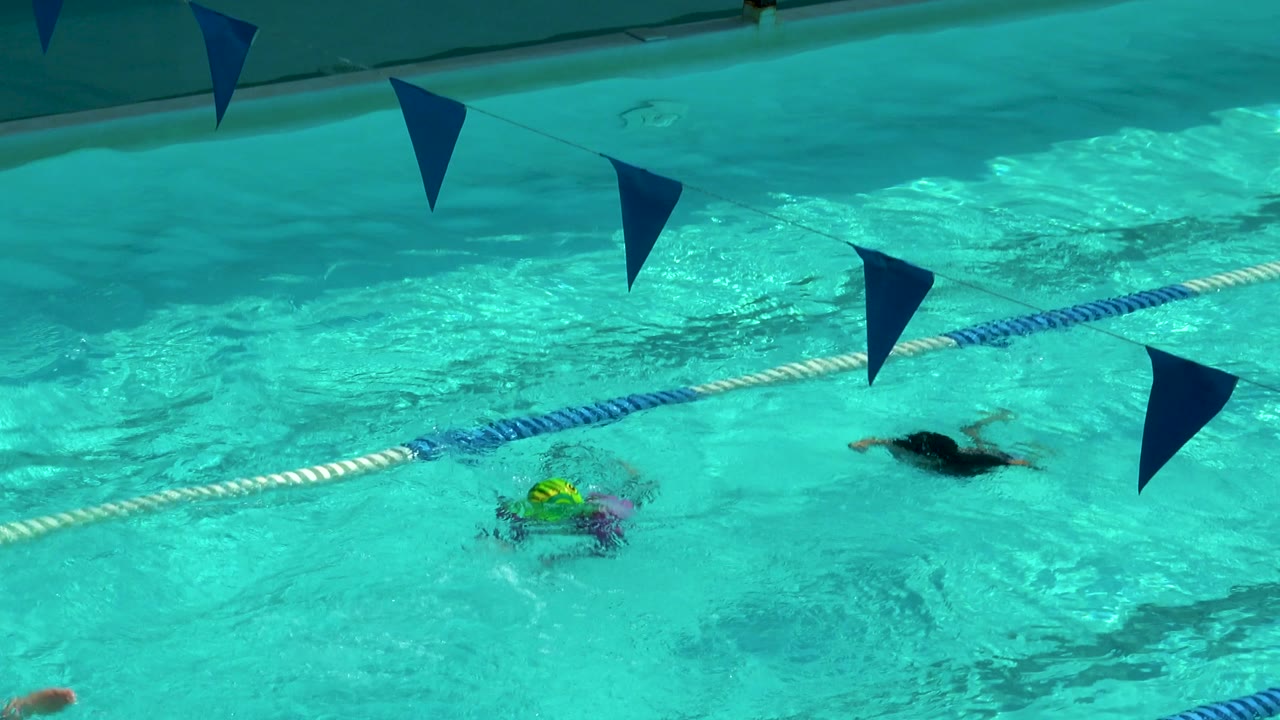 Children swim across turquoise pool lanes in Cabo San Lucas, with blue pennant flags overhead and colorful caps visible near the lane rope.