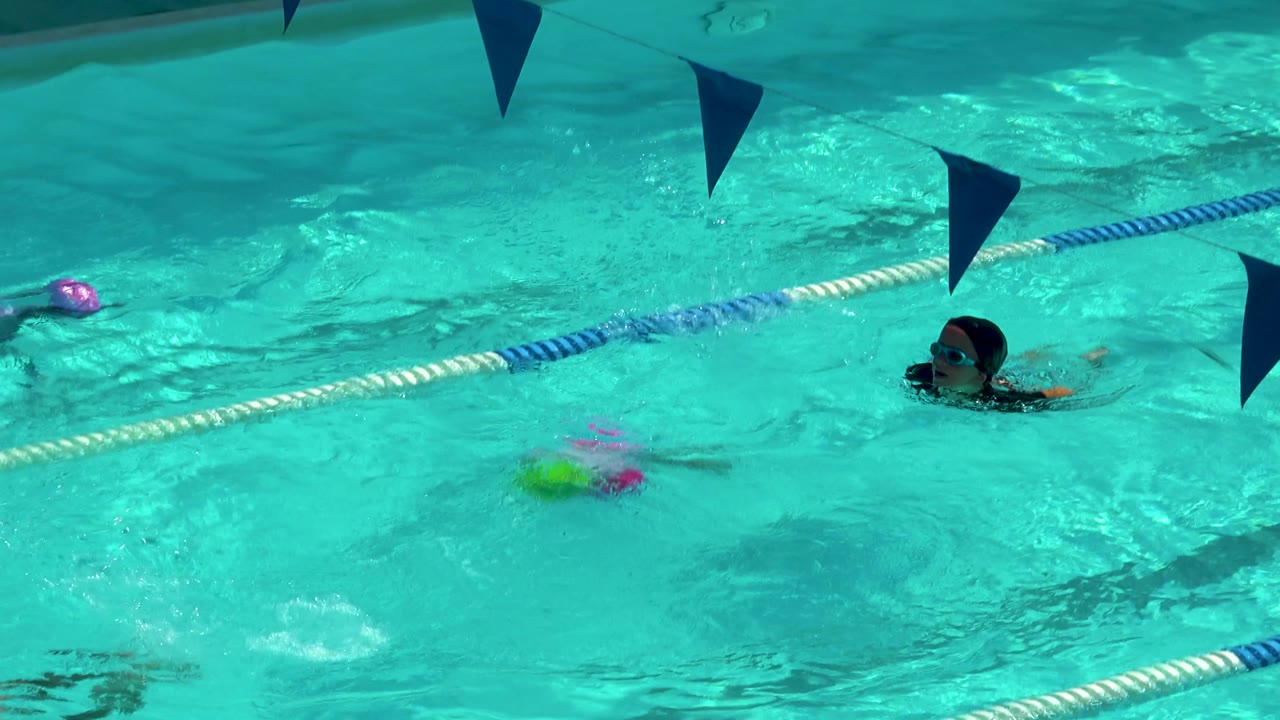 Several children swim through turquoise lanes in Cabo San Lucas, including a pink-capped swimmer near the top and a goggled swimmer at the right edge.