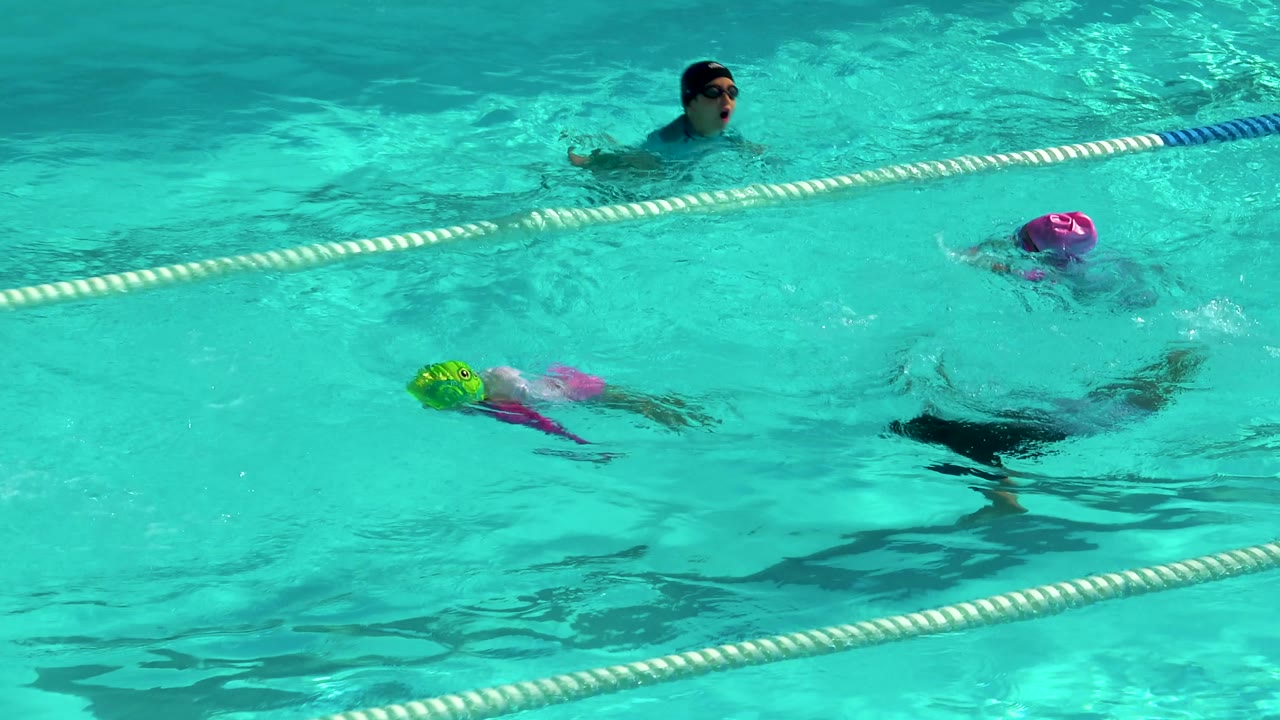 Children swim through turquoise lanes in a Cabo San Lucas pool while an adult swimmer is visible near the upper lane at the start of the clip.
