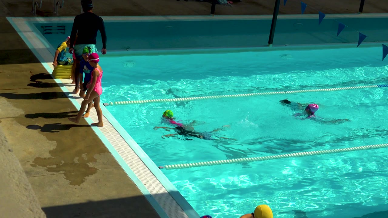 At a Cabo San Lucas outdoor pool, children in bright swim caps gather near the deck while two swimmers move through turquoise water beside a lane rope.