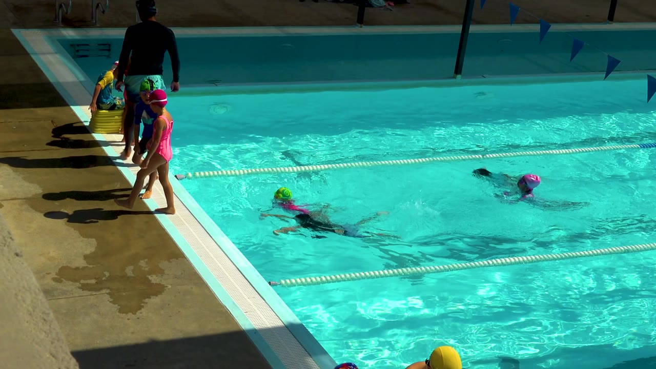 Swimmers cross the pool while children wait near an instructor on deck.