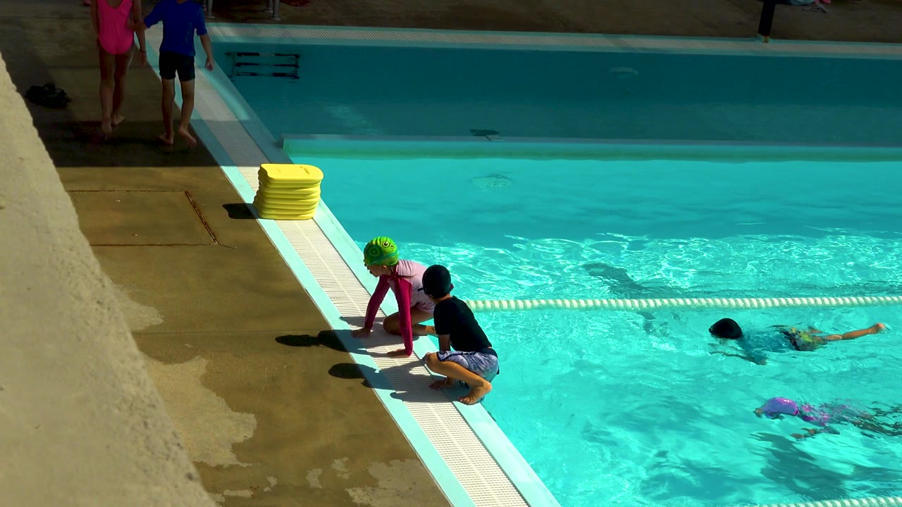 Children in swim caps gather at the edge of a bright outdoor pool in Cabo San Lucas, Mexico, with yellow kickboards on the deck and swimmers in the water.