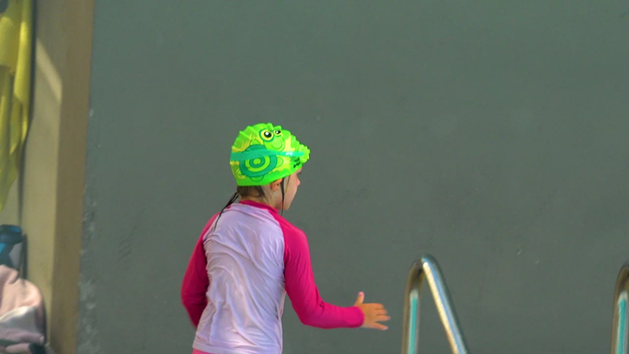 A child in a bright green swim cap and pink rash guard faces away near a metal pool rail in Cabo San Lucas, Mexico.