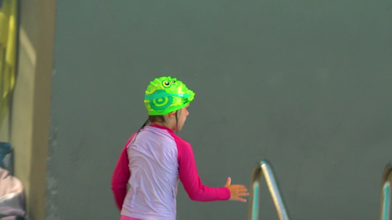 Child in green swim cap seen from behind near a pool rail.