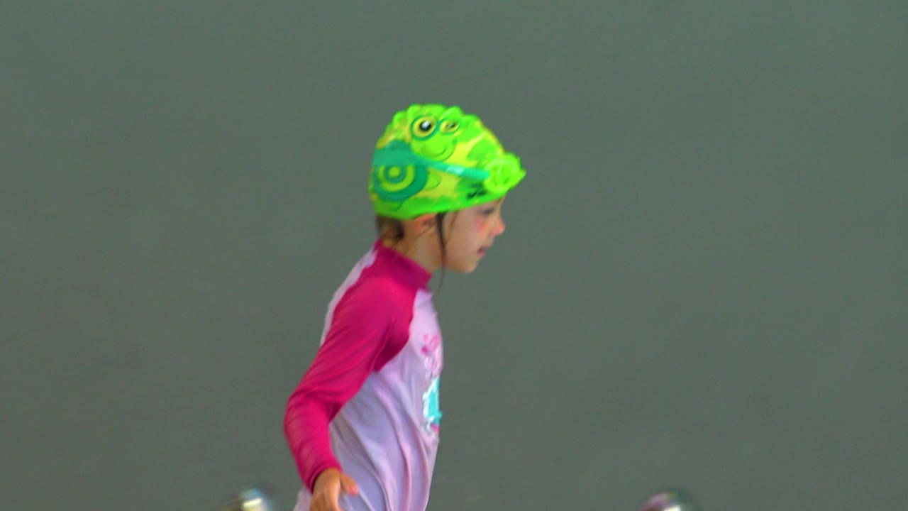 A child in a bright green swim cap and pink swim shirt is shown in side profile against the plain green pool wall in Cabo San Lucas, Mexico.