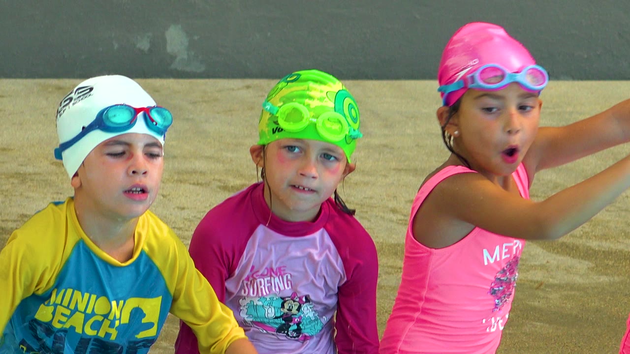 Three children in Cabo San Lucas wear colorful swim caps, goggles, and rash guards near a wet concrete poolside wall.