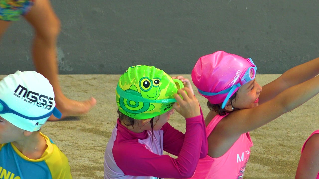 Children in colorful swim caps and bright swim shirts gather beside a wet concrete surface in Cabo San Lucas, Mexico, with faces partly turned away at the start of the clip.