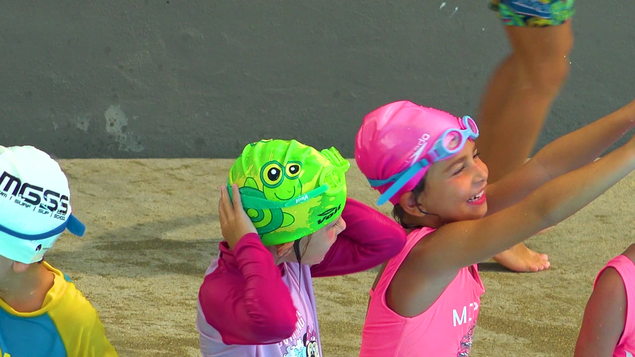 The girl in the pink swim cap smiles and points upward with both hands while two nearby children in swim caps look on in Cabo San Lucas, Mexico.