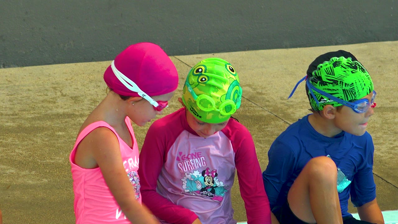 Three children in Cabo San Lucas sit close together at a pool edge, with a pink swim cap on the left, a green cap in the middle, and a blue-shirted child on the right.
