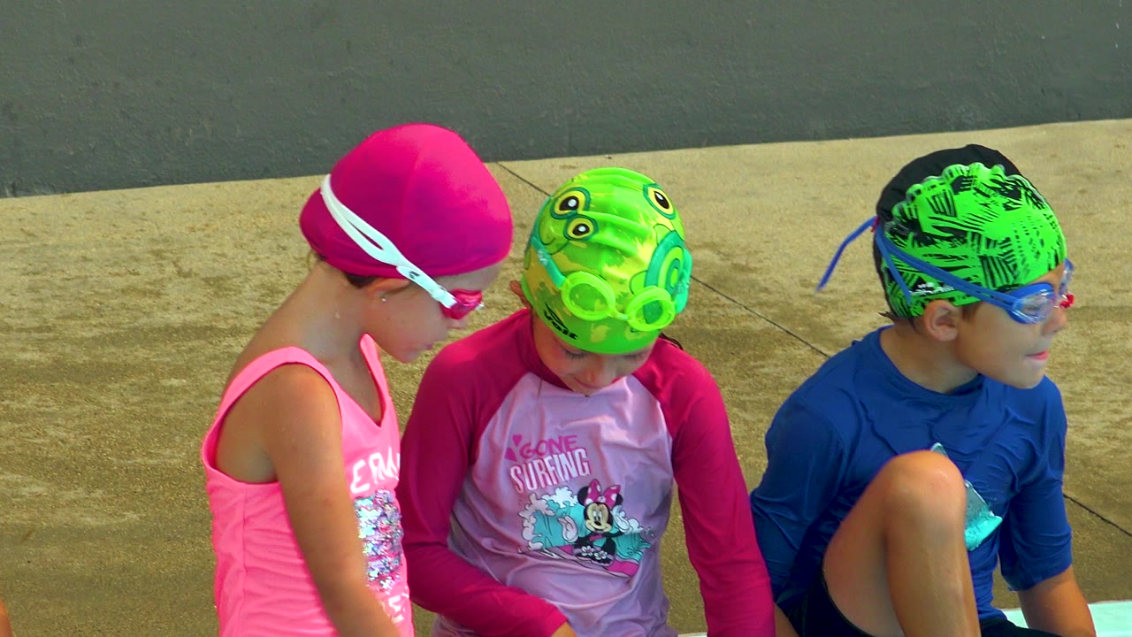 Three children in goggles sit beside the pool on a concrete deck.