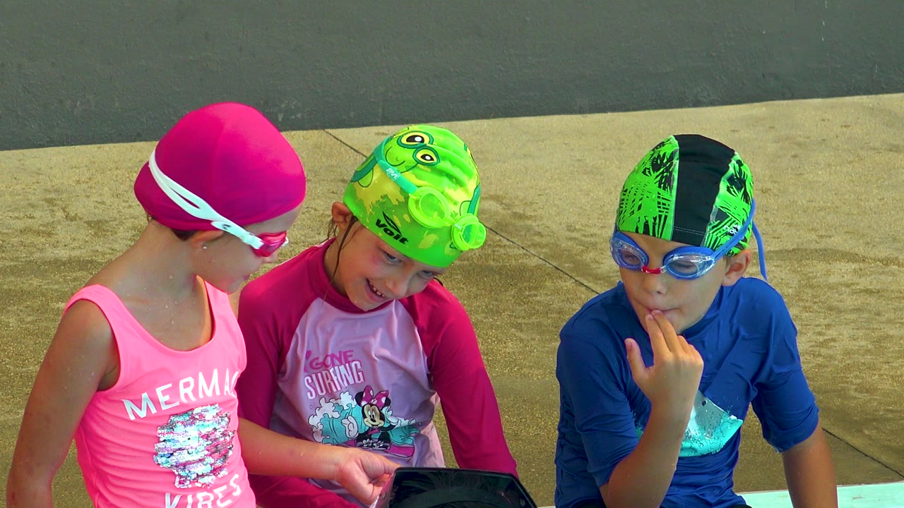 Three children sit close together beside a pool in Cabo San Lucas, Mexico, with a pink-capped child looking toward two children in green and blue swim caps.