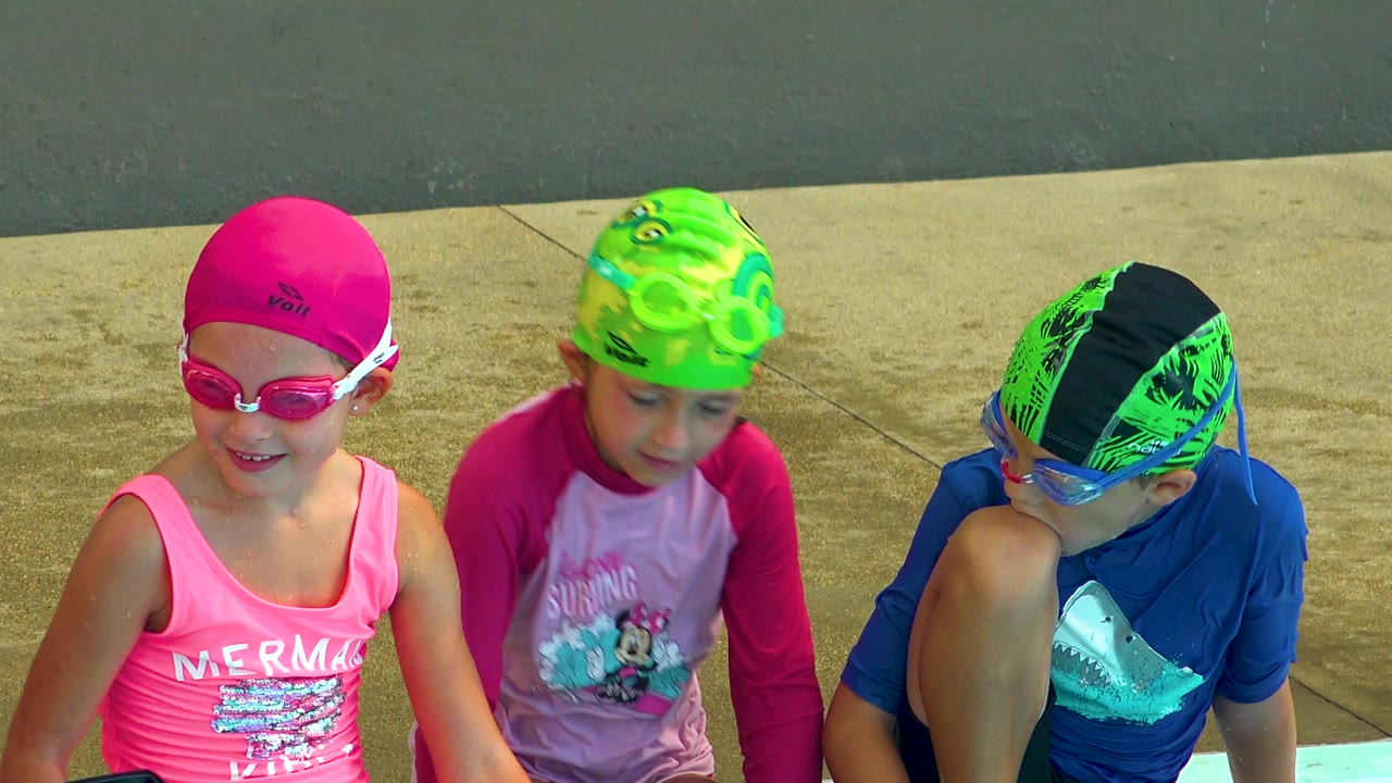 Three children in bright swim caps and goggles sit close together at a pool edge in Cabo San Lucas, Mexico, with a sunlit concrete deck behind them.