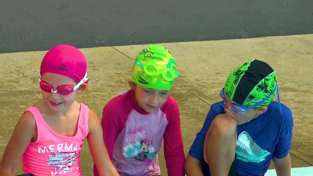Three children sit poolside while the child in the pink cap smiles forward.
