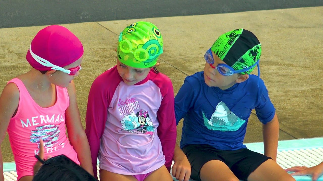 Three young children sit above a pool in Cabo San Lucas, with a girl in a pink swim cap looking toward two children in green swim caps and goggles.