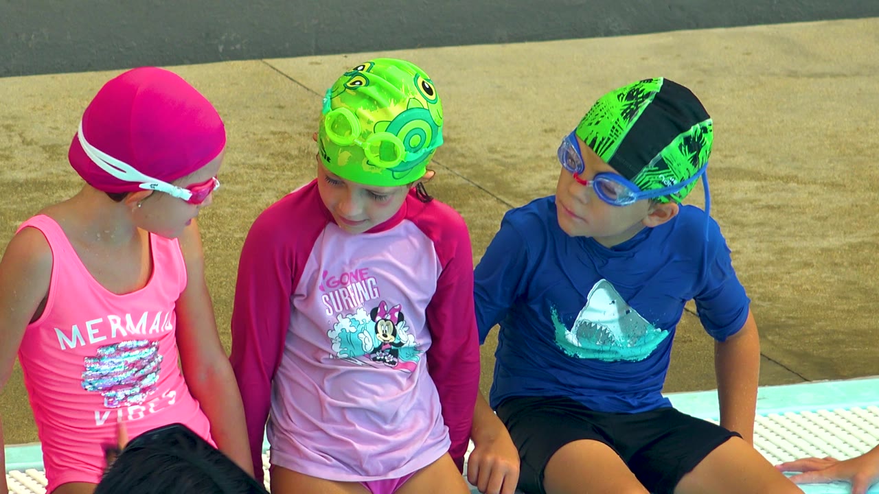 Three children sit close together at the pool edge in swimwear.
