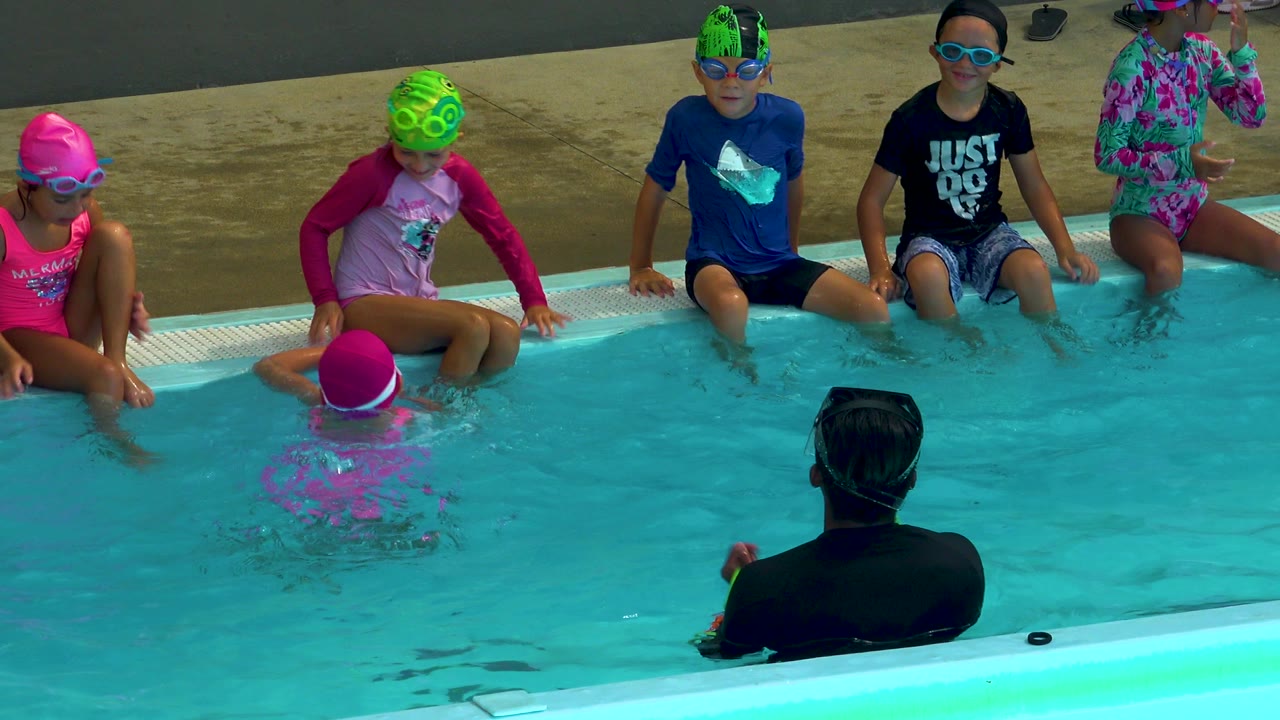 Children wearing swim caps and goggles sit along a pool edge in Cabo San Lucas, Mexico, while an adult instructor faces them from the foreground water.