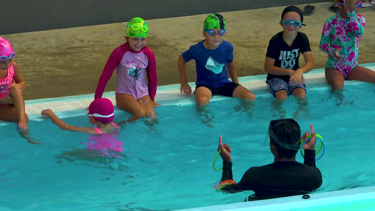 An adult instructor in the foreground water gestures toward children in swim caps seated on the pool edge in Cabo San Lucas, Mexico.