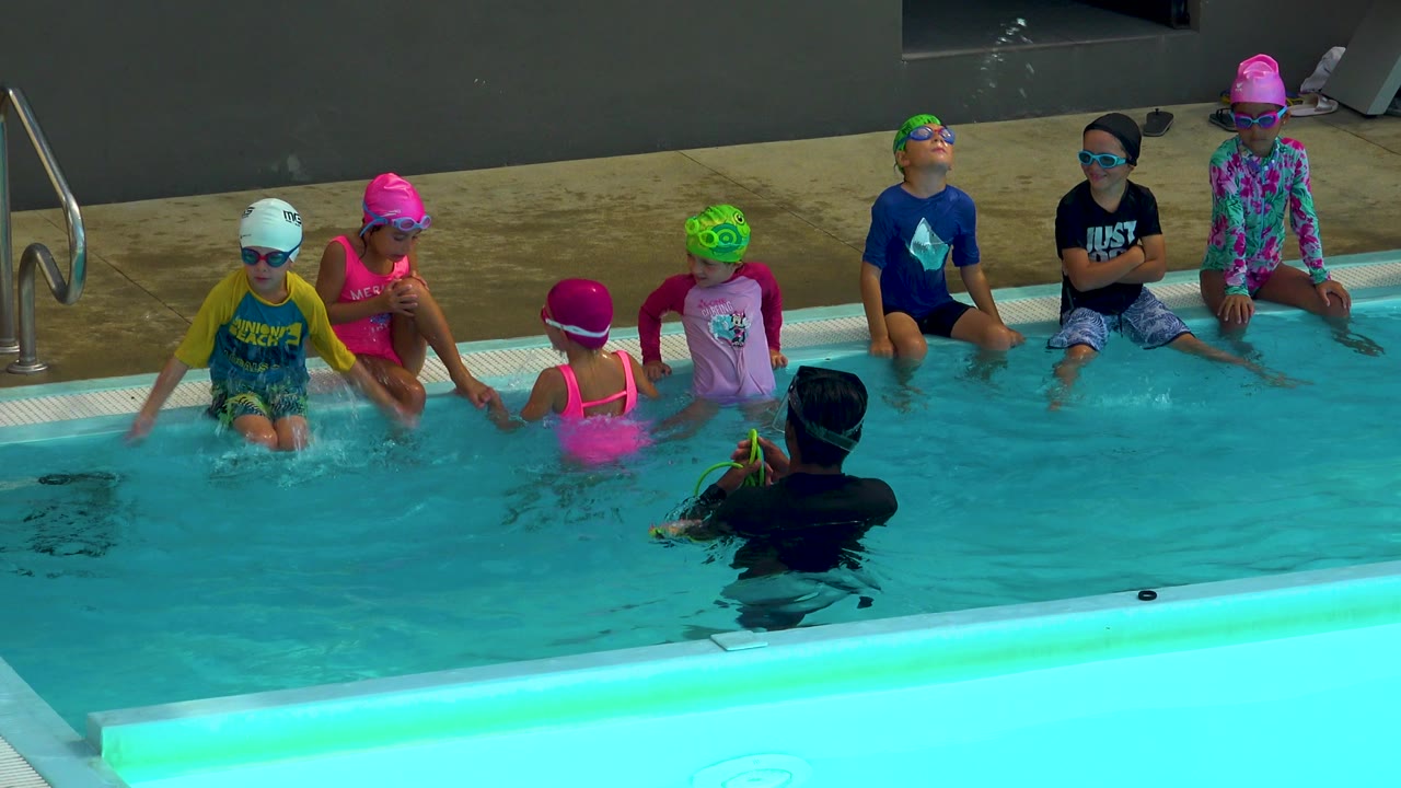 Children in colorful swim caps and goggles sit along a pool edge in Cabo San Lucas while an adult instructor faces them from the water.