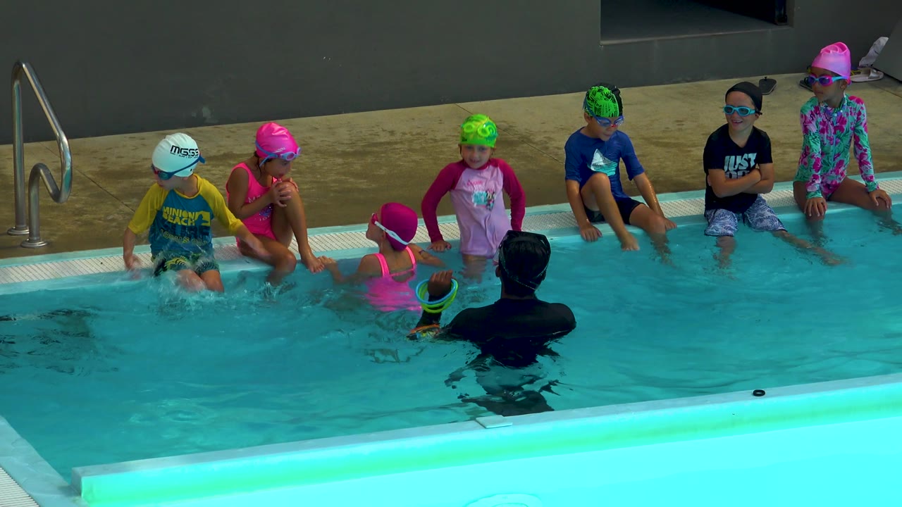 Children in pink, green, white, and dark swim caps sit along the Cabo San Lucas pool edge as the instructor faces them from the foreground water.