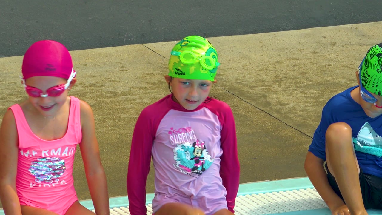 At the Cabo San Lucas pool edge, the girl in pink swimwear faces forward while another child in a green cap looks ahead and a child in blue is cropped at the side.