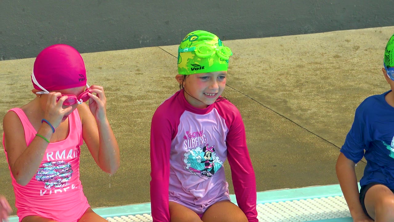 Children in bright swim caps and goggles sit along a pool edge in Cabo San Lucas, Mexico, with turquoise water and wet concrete visible.