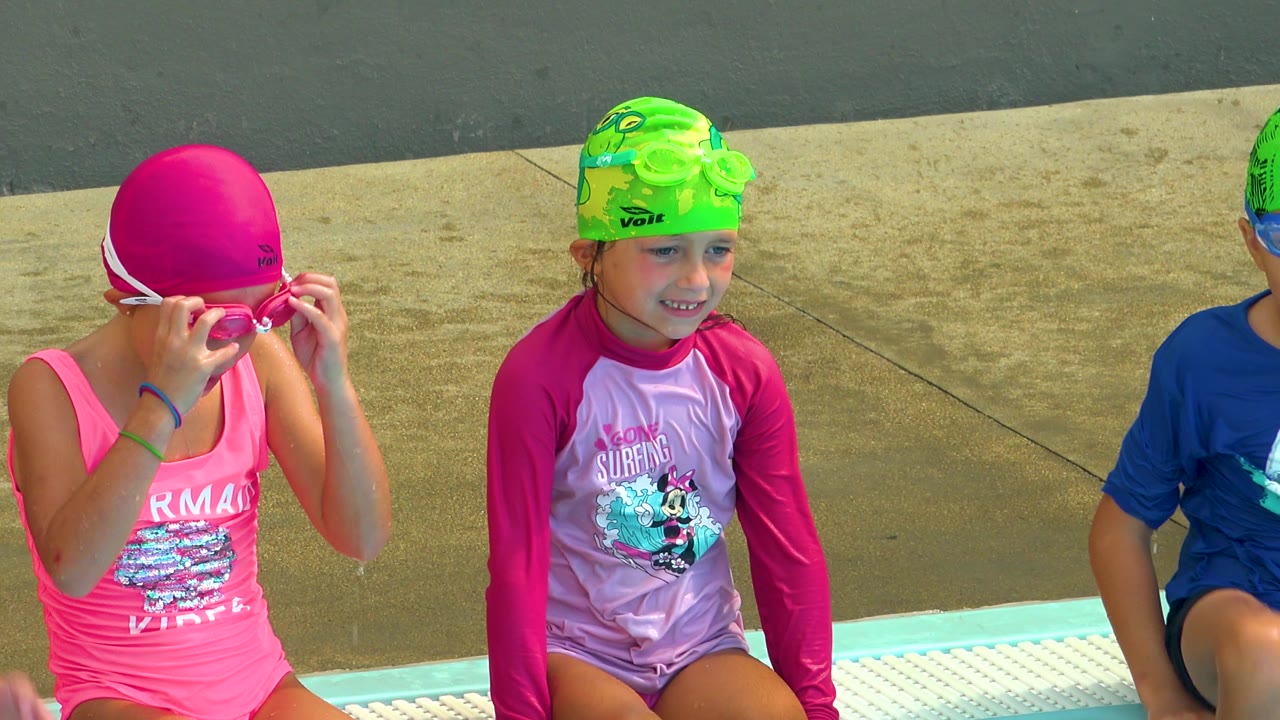 A girl adjusts pink goggles while other children sit beside her at the pool.