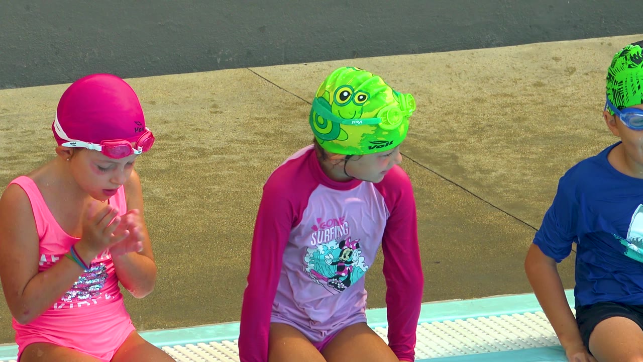 Children sit in a row at the pool edge in Cabo San Lucas, with a pink-capped child leaning forward and a green-capped child looking down.
