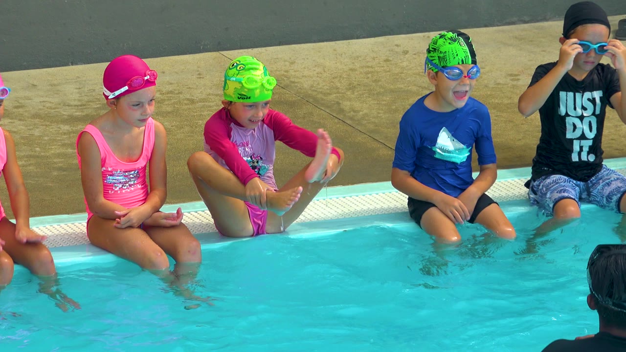 At a Cabo San Lucas pool, several children in bright swim caps and goggles sit along the edge while an adult instructor is partly visible from behind in the water.